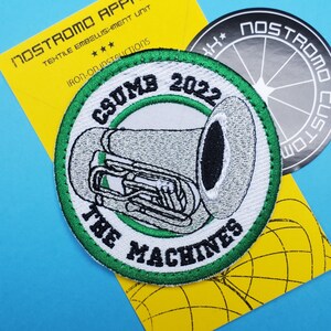 Pode incluir: Uma coleção de patches e adesivos. O patch principal é redondo com uma borda verde e apresenta uma tuba e o texto "CSUMB 2022 THE MACHINES". Um cartão amarelo com "NOSTROMO APPAREL" está atrás do patch. Há também um adesivo redondo com "NOSTROMO CUSTOM".