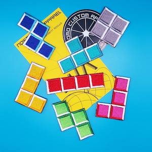 May include: A set of ten colorful iron-on patches featuring Tetris game pieces. The patches are in various colors, including yellow, blue, red, green, and purple. The patches are arranged in a random pattern on a blue background. The patches are all different sizes and shapes, and they are all made of a durable fabric.
