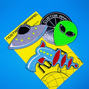 May include: A set of four iron-on patches featuring a green alien face, a blue and red space ray gun, a gray UFO, and a black and white logo with the text "Custom Apparel".