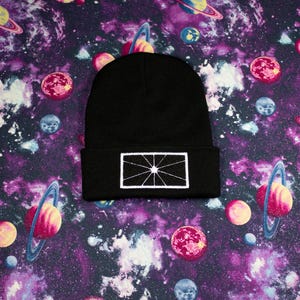 Puede incluir: Un gorro negro con un diseño geométrico blanco bordado. El gorro se encuentra sobre un vibrante fondo morado y azul con temática espacial, con planetas y galaxias.