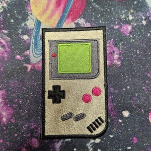 May include: An embroidered patch shaped like a handheld video game console. The device is light gray with a bright green screen, a black directional pad, two pink circular buttons, two small gray buttons, and black speaker slits.