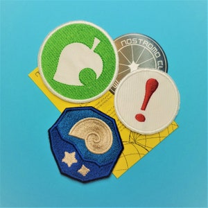 May include: A collection of embroidered patches featuring various designs. One patch has a green circle with a white leaf, another has a red exclamation point, and a third has a blue and gold seashell design. A silver patch reads "NOSTROMO CLUB".