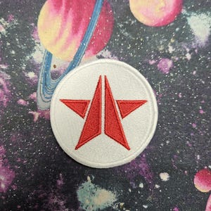 May include: A circular embroidered patch featuring a white background with a red star-like emblem in the center. The emblem consists of two symmetrical, sharp-pointed red shapes that form a stylized star design with a vertical split.