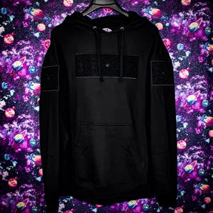 May include: Black hooded sweatshirt with a front pocket and a star emblem. The sweatshirt has a rectangular patch on the chest and smaller patches on the sleeves. The background is a galaxy print.