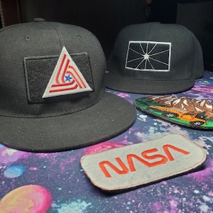 May include: Two black baseball caps with embroidered patches. One cap has a red, white and blue triangle patch with a star. The other cap has a black and white patch with a starburst design. A grey patch with the word "NASA" in red lettering is on a purple and black background.