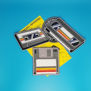 Pode incluir: Três patches bordados com tecnologia retro: uma fita cassete, um disquete e uma fita VHS. Cada patch tem um design de listras amarelas, vermelhas e laranja.