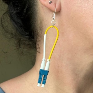 May include: A pair of unique earrings featuring a yellow and white cable design with blue accents. The earrings hang from silver hooks and are worn on the ear. The design is modern and eye-catching.