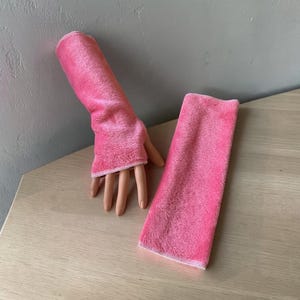 May include: A pair of bright pink fingerless gloves made of a soft, plush, fuzzy fabric. One glove is worn on a mannequin hand, while the other lies flat next to it. Both gloves feature a simple, straight-cut design with finished edges.