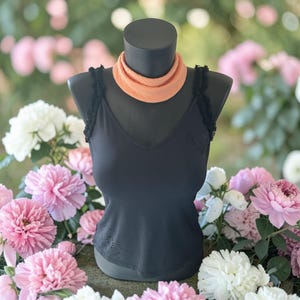 May include: A black tank top with a V-neck and delicate lace trim on the straps. The top is displayed on a mannequin, paired with an orange scarf. The background features a variety of pink and white flowers.