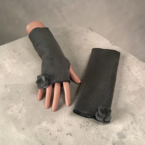 Peut inclure: Une paire de gants sans doigts en polaire texturée gris foncé. Chaque gant présente une petite fleur en tissu assortie ornant la zone du poignet. Un gant est présenté sur un présentoir en forme de main, tandis que l'autre est posé à plat.
