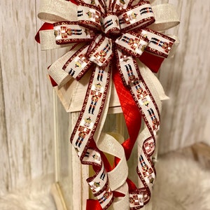 May include: A white lantern with a red and white nutcracker ribbon bow. The bow is tied around the top of the lantern and the ribbon tails hang down the sides. The lantern is sitting on a white fluffy surface.