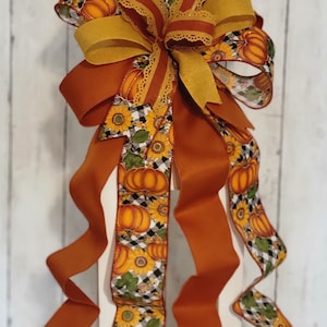 May include: A decorative fall-themed bow featuring a combination of solid orange and patterned ribbons. The patterned ribbons display pumpkins, sunflowers, and a black and white checkered design. The bow is suitable for seasonal decor.