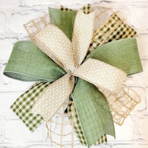 May include: A green and beige bow with a burlap mesh base. The bow is made of three different types of ribbon: a solid green ribbon, a beige ribbon with white polka dots, and a green and white gingham ribbon.