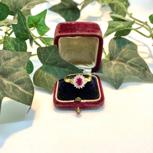 Puede incluir: Un anillo de oro con una piedra ovalada roja rodeada de piedras blancas más pequeñas. El anillo está en una caja de anillo de terciopelo rojo.