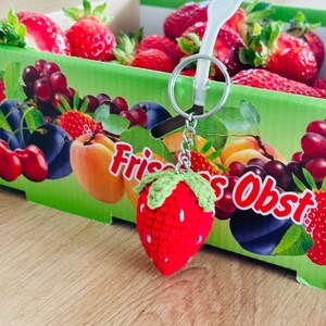 May include: A red crocheted strawberry keychain with green leaves hangs from a silver chain. The keychain is in front of a box of fresh fruit, including strawberries, grapes, and plums. The box is labeled "Frisches Obst".