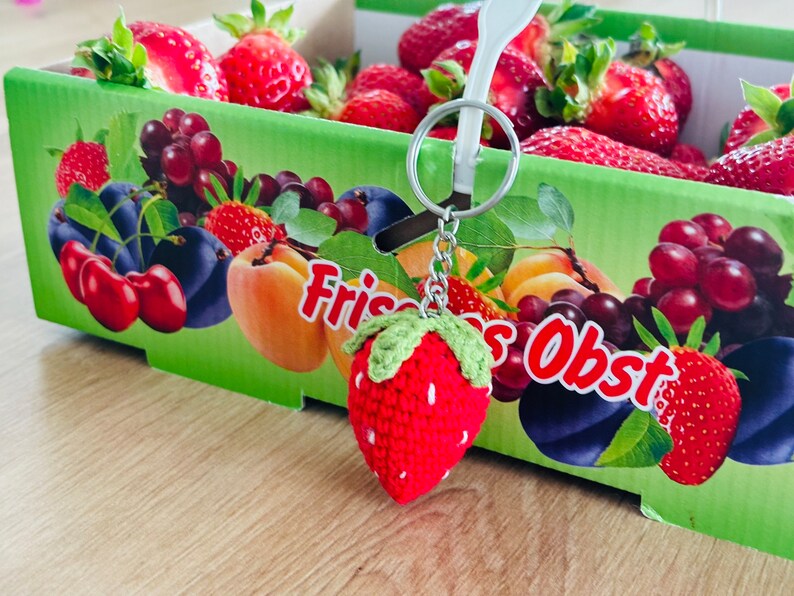 May include: A red crocheted strawberry keychain with green leaves hangs from a silver chain. The keychain is in front of a box of fresh fruit, including strawberries, grapes, and plums. The box is labeled "Frisches Obst".
