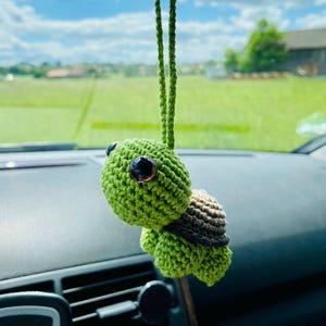 Könnte beinhalten: Eine grüne gehäkelte Schildkröte, die an einem Rückspiegel in einem Auto hängt. Die Schildkröte hat schwarze Augen und einen braunen Panzer.