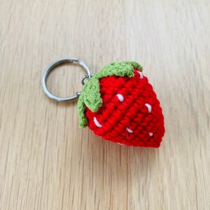 May include: A red crocheted strawberry keychain with green leaves and white seeds. The strawberry is attached to a silver key ring.