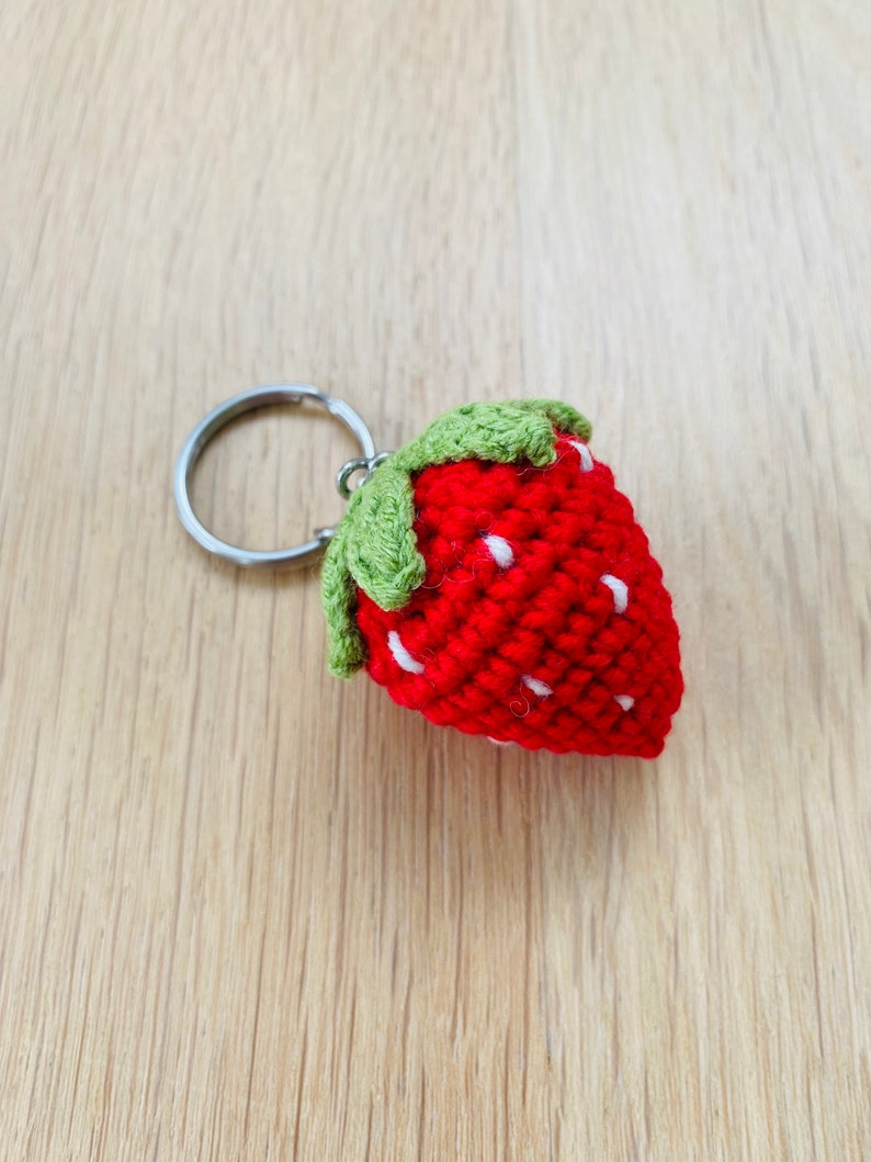 May include: A red crocheted strawberry keychain with green leaves and white seeds. The strawberry is attached to a silver key ring.