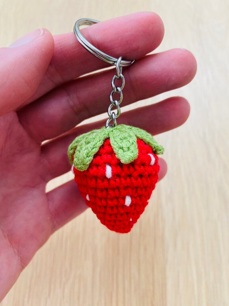 May include: A red and green crocheted strawberry keychain with white accents. The strawberry is attached to a silver key ring.