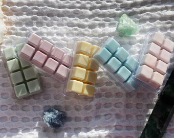Op de afbeelding: Assortiment van negen wax melts in vier verschillende kleuren: groen, roze, blauw en geel. De wax melts zitten in een transparante plastic verpakking en zijn gerangschikt in een rasterpatroon.