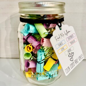 Op de afbeelding: Een heldere Mason jar gevuld met kleurrijk opgerold papier, vastgebonden met een zwart lint en afgedekt met een zilveren deksel. Een wit label leest "Read Me When..." en "Color Coded Bible Verses."