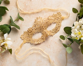 May include: A gold lace masquerade mask with a delicate design and rhinestones. The mask is tied with a gold ribbon.
