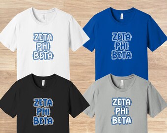 May include: Four t-shirts in different colors, white, blue, black, and gray, with the text "Zeta Phi Beta" printed in blue on the front.