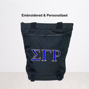 May include: Black tote bag with a zippered pocket and adjustable straps. The bag is embroidered with the Greek letters "ΣΓΡ" in blue and gold.