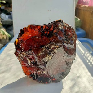 May include: A large, irregularly shaped piece of dark amber with swirling patterns and inclusions. The amber is translucent and has a reddish-brown color.
