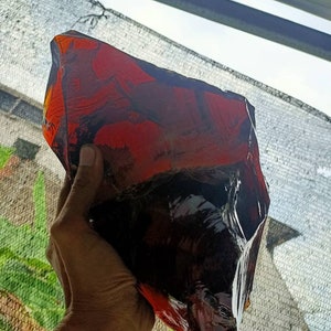 May include: A large, irregularly shaped piece of red obsidian, a type of volcanic glass, with a dark, almost black, interior. The obsidian is being held in a hand.