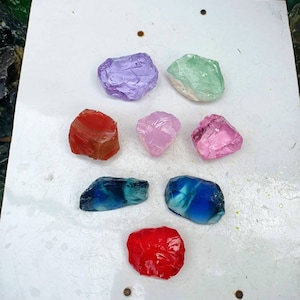 Op de afbeelding: Assortiment van acht kleurrijke glazen stenen in verschillende vormen en maten. De stenen zijn gerangschikt op een wit oppervlak. De kleuren omvatten rood, roze, paars, blauw en groen.
