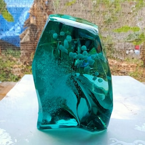 May include: A teal blue glass sculpture with white bubbles trapped inside. The sculpture has an irregular shape and is sitting on a white surface.