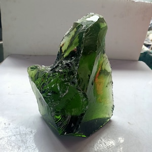 Puede incluir: Una pieza grande de vidrio verde de forma irregular con una superficie rugosa y texturizada. El vidrio es translúcido y tiene un ligero tono verdoso.