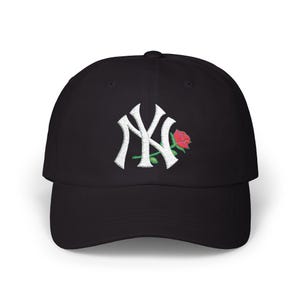 Puede incluir: Gorra de béisbol negra con el logotipo "NY" bordado en blanco y una rosa roja. La gorra tiene una visera curva y una correa ajustable. Un accesorio de moda para el uso diario.