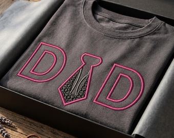 May include: A charcoal gray crew neck t-shirt featuring the embroidered text DAD in pink, with the letter A replaced by a black and gray patterned necktie graphic. The shirt is folded inside a black gift box with gray tissue paper.