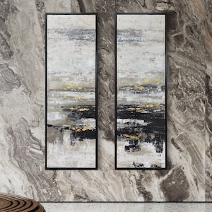 May include: Two abstract paintings in black frames, each featuring a white background with black and gold brushstrokes. The paintings are hung on a gray marble wall.