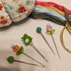 May include: Five colorful glass needle threaders with different designs, including a frog, a flower, and a pearl. The threaders are on a white fabric background.