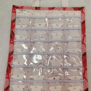 May include: A red and white fabric thread organizer with a white handle. The organizer has clear plastic pockets for holding spools of thread. The fabric has a floral pattern. The edges are trimmed with a red and white floral fabric.