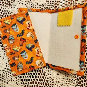 May include: A Halloween-themed fabric wallet with a white interior and a yellow felt pocket. The front of the wallet features a repeating pattern of orange, black, and white masks and characters.