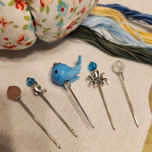 May include: Five decorative pins with blue and white glass animal figures. The pins feature a whale, an octopus, and a dolphin. The pins are on a white surface.