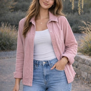 Puede incluir: Una camisa de pana rosa claro con un bolsillo delantero, usada sobre una camiseta sin mangas blanca y combinada con vaqueros azul claro. El conjunto se completa con un collar y una pulsera dorados. La persona está al aire libre.