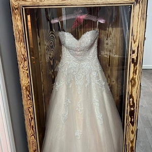 May include: A white wedding dress with lace detailing is displayed in a wooden display case with a glass front. The case has a dark wood finish and is decorated with the letters "R" and "J" on top.