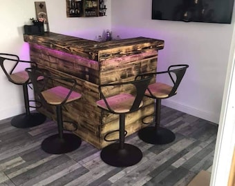 May include: A corner bar made from reclaimed wood with a black metal frame and three black bar stools. The bar has a pink LED light strip underneath the countertop. The wall behind the bar has a wooden shelf with the text "Nobody gets out sober".