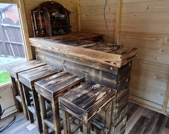 May include: A rustic wooden bar with a burnt finish and four matching stools. The bar has a shelf above the counter and is made of reclaimed wood.