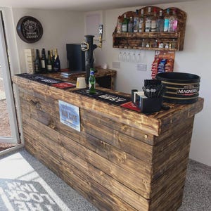 May include: A wooden home bar with a dark stain, featuring a tap, bottles, and a "Man Cave" sign. The bar has a built-in shelf for liquor bottles and a bucket labeled "Magners". A Jack Daniel's sign hangs on the wall.