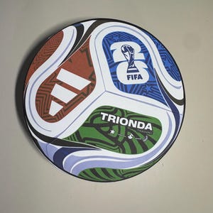 May include: A round soccer ball featuring a multi-colored panel design with white, red, blue, and green sections. It displays the Adidas logo, the FIFA 26 logo, and the text TRIONDA TRAINING with small star icons printed on the green panel surface.