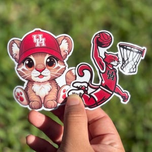 May include: Two cartoon stickers featuring a red and white mascot. The first sticker shows a cartoon cougar wearing a red baseball cap with a white "UH" logo. The second sticker shows a cartoon cougar dunking a basketball. Both stickers have a white outline.