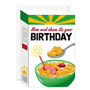 May include: A rectangular greeting card featuring a cereal box design with a green bowl of cereal, strawberries, and a candle. The text reads, Rise and shine it's your BIRTHDAY. The design includes a yellow sunburst graphic and a silver spoon.