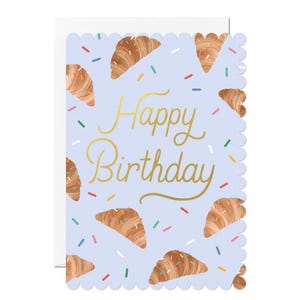 May include: A light blue birthday card with a scalloped edge. The card features gold script that reads "Happy Birthday" and illustrations of croissants with colorful sprinkles. The card is blank inside and comes with a white envelope.